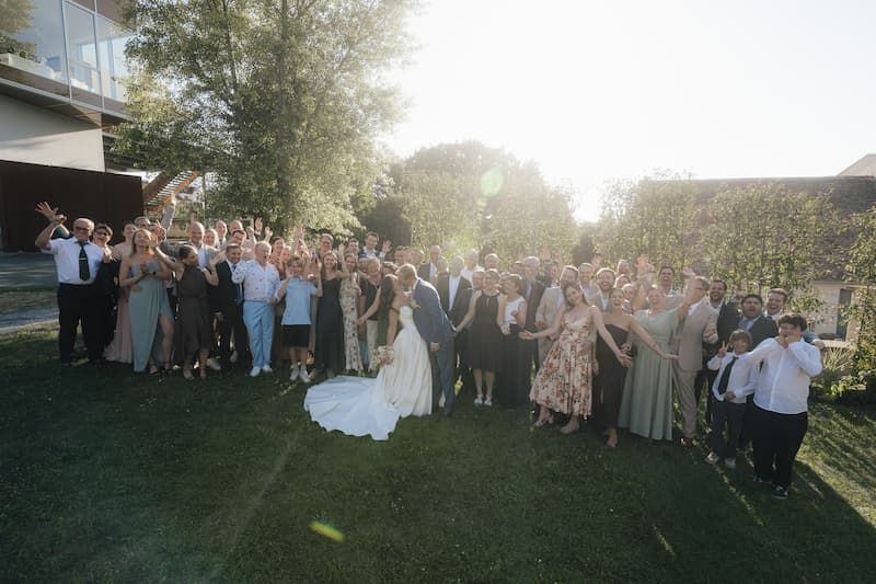 Large wedding group photo with bride and groom surrounded by cheering guests on a sunny lawn.