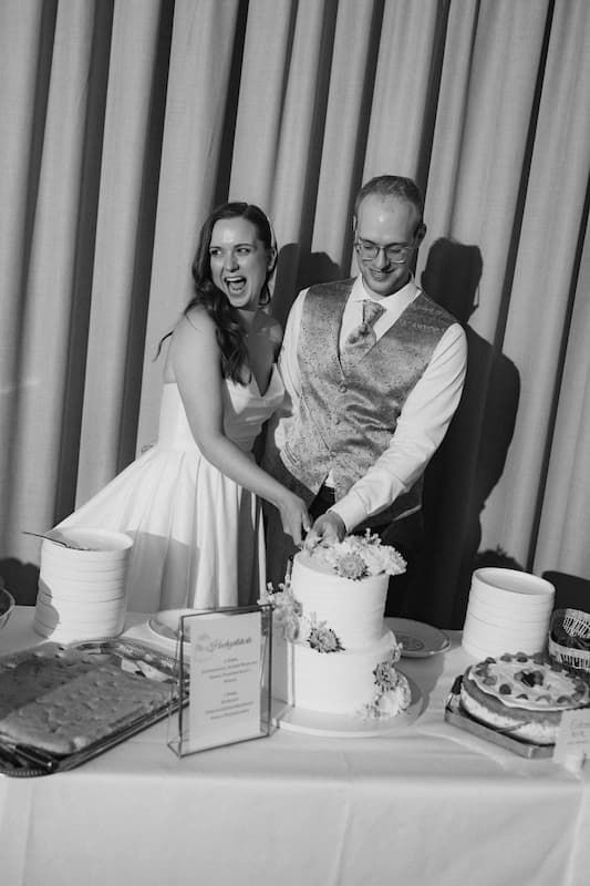 Bride and groom cutting their wedding cake together at reception table in black and white photo.
