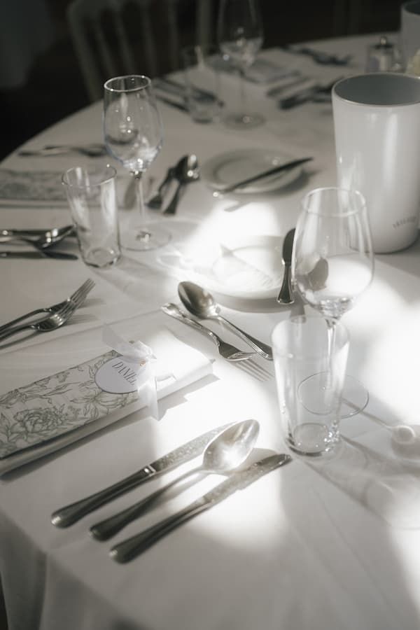 Elegant white table setting with silverware, wine glasses, and menu cards in soft natural light.