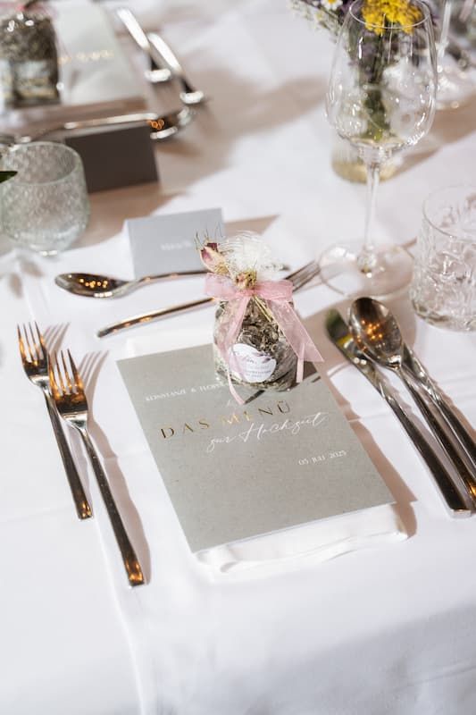 Elegant wedding table setting with gray menu card, silver cutlery, crystal glasses, and a small floral jar favor.