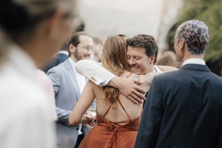 Guests embracing warmly at an outdoor wedding reception, smiling man hugging woman in backless rust dress.