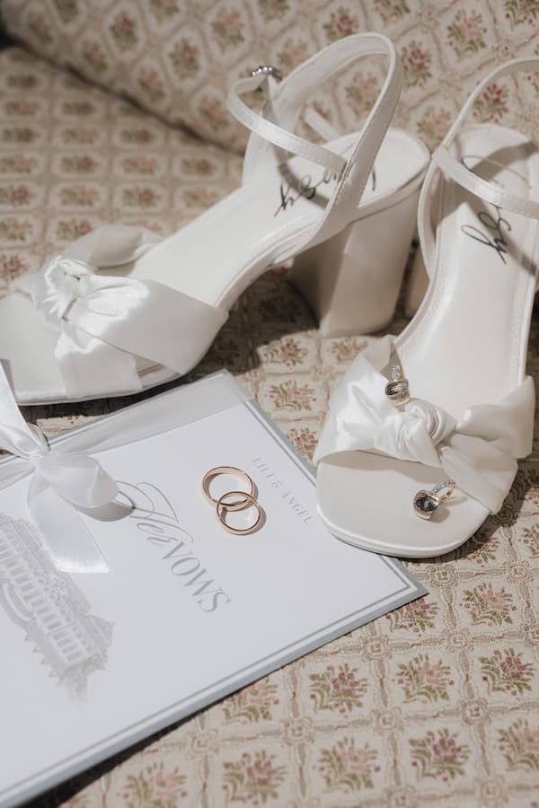 White bridal heels with satin bows beside gold wedding rings on a vows booklet, elegant wedding accessories flat lay.