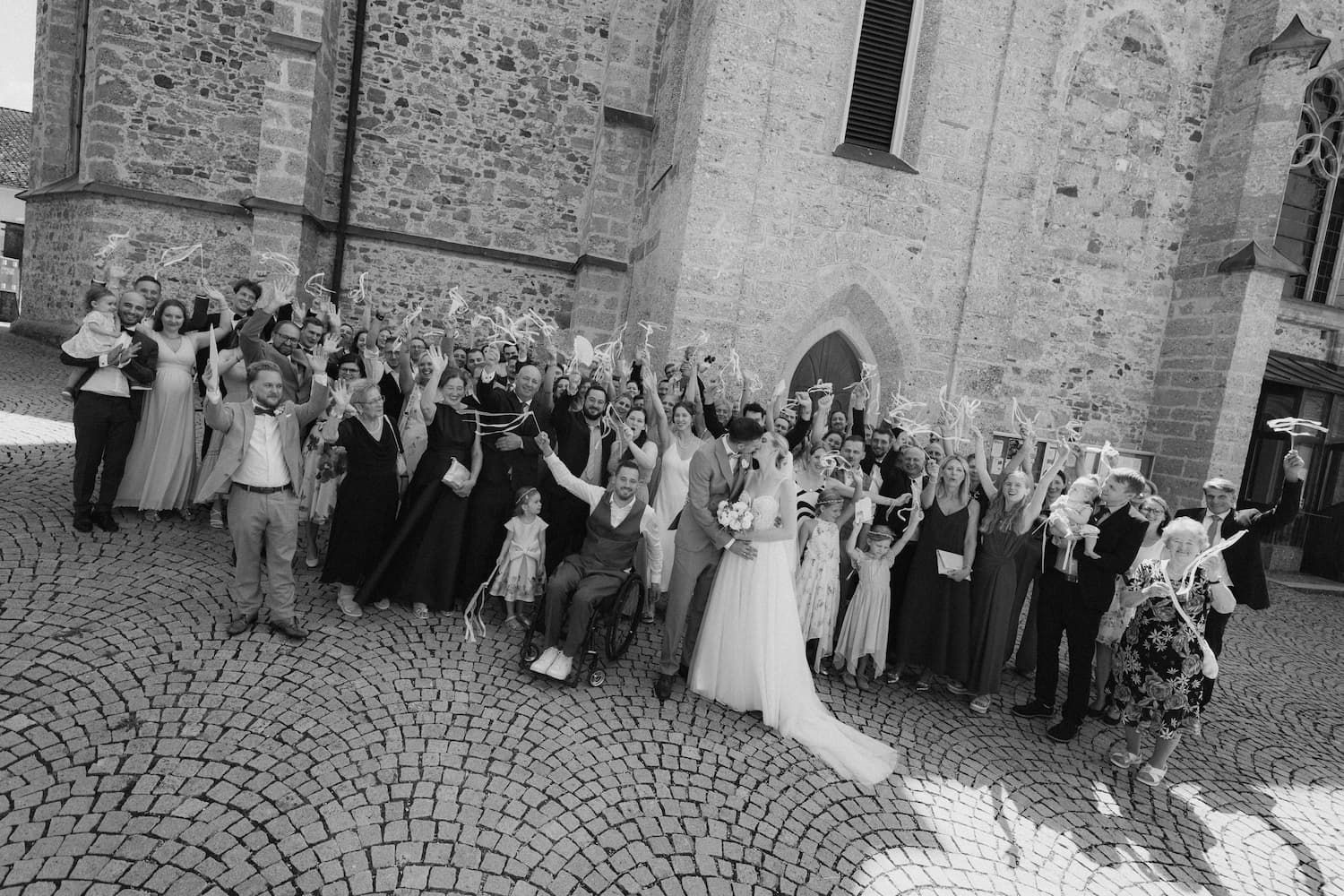 Black and white wedding photo of bride, groom, and guests cheering outside a historic stone church.