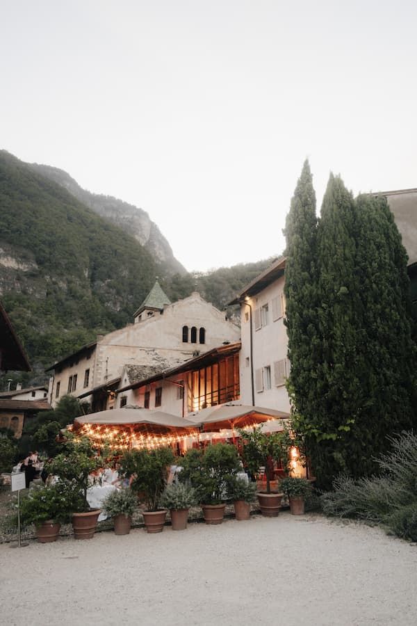 Charming European building with warm string lights, cypress trees, and mountain backdrop at dusk.