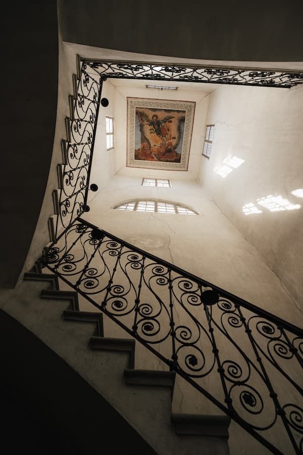 Elegant spiral staircase with ornate wrought iron railings leading to a ceiling fresco in a historic building.