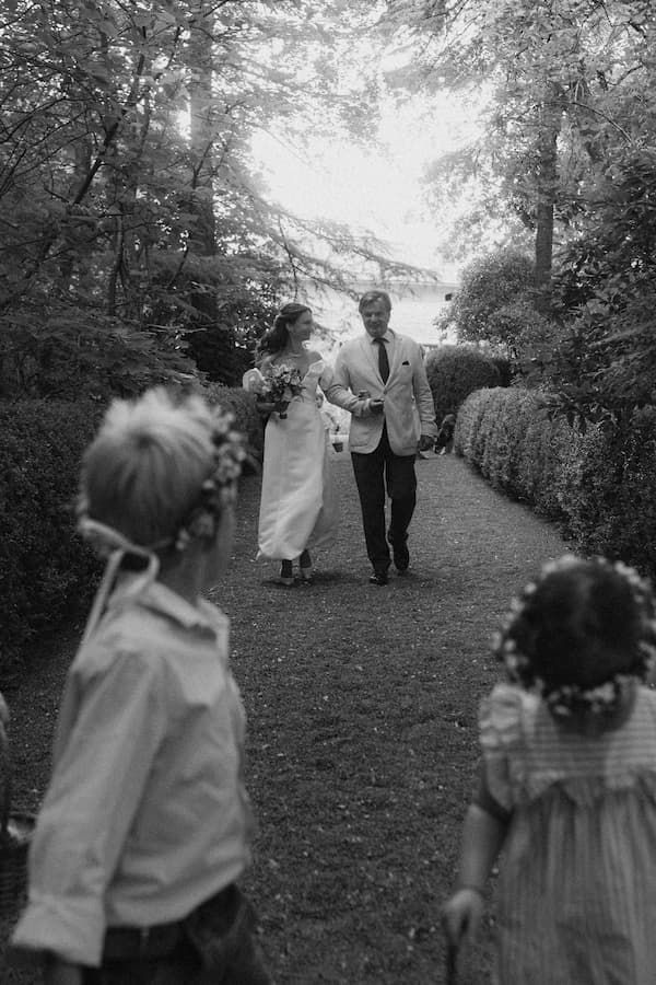 Black and white photo of a bride and groom walking down a garden path, with two flower girl children in the foreground.