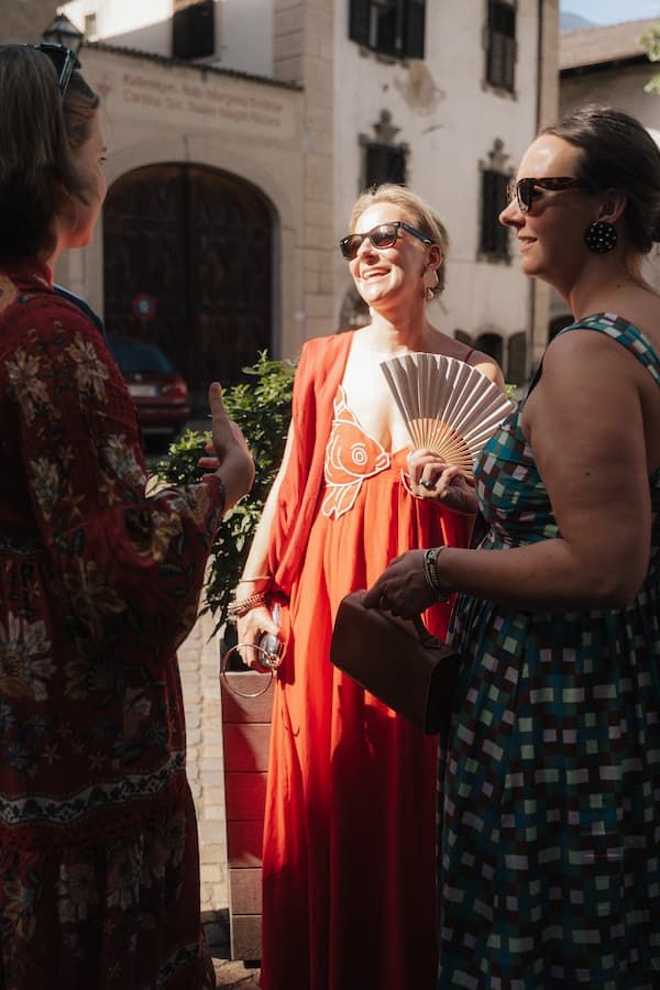 Three stylish women in summer dresses chat outdoors, one in a red gown holding a decorative fan.