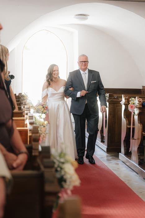 Bride walks down church aisle with father on red carpet, smiling joyfully during wedding ceremony.