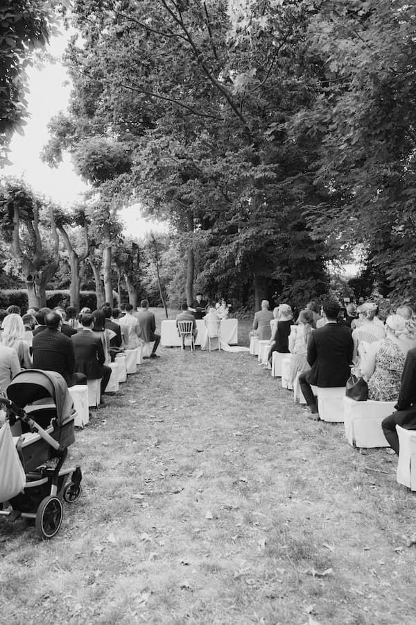 Black and white photo of an outdoor wedding ceremony with guests seated in rows along a tree-lined garden aisle.