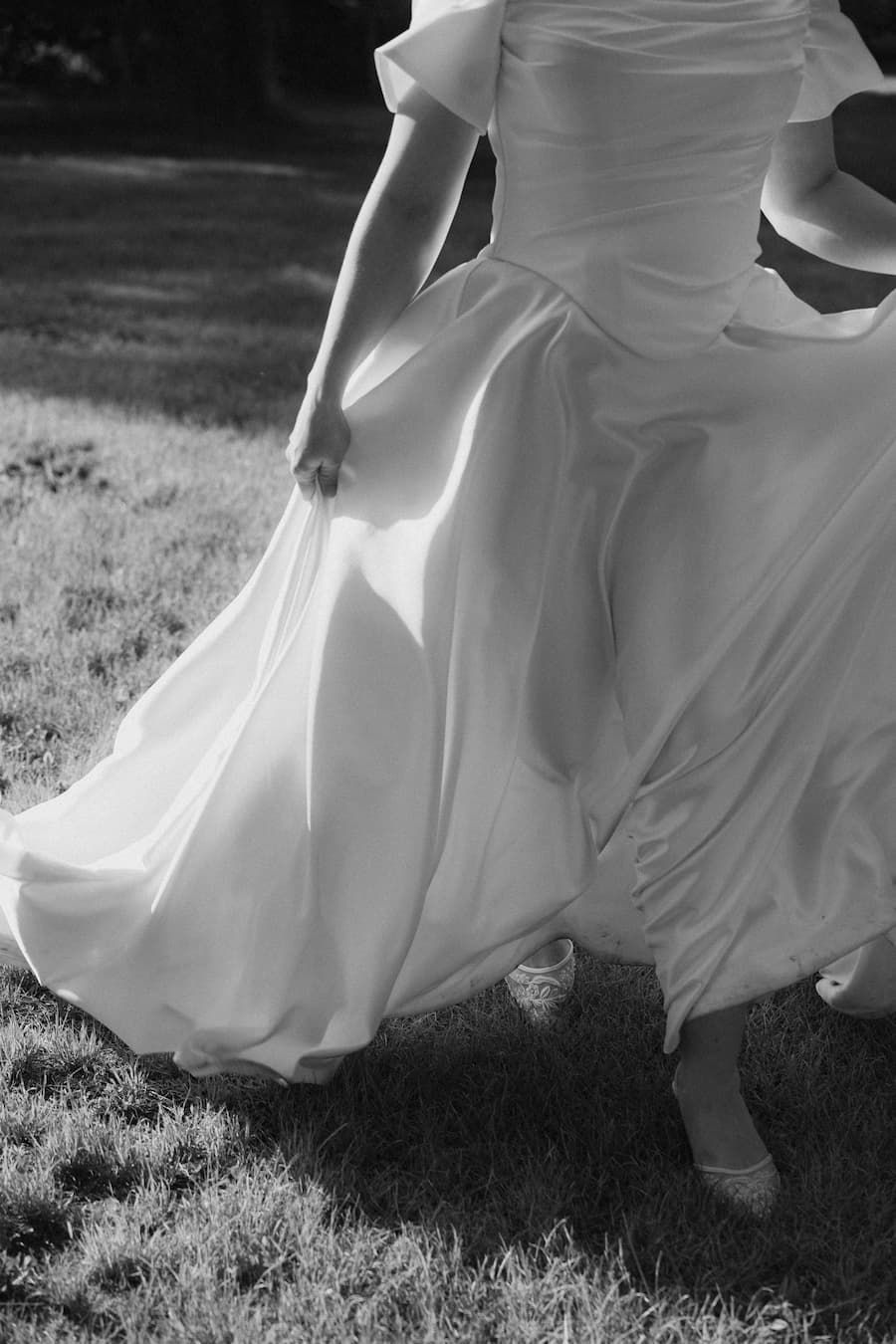 Black and white photo of a bride in a flowing white off-shoulder gown holding her dress while walking on grass.