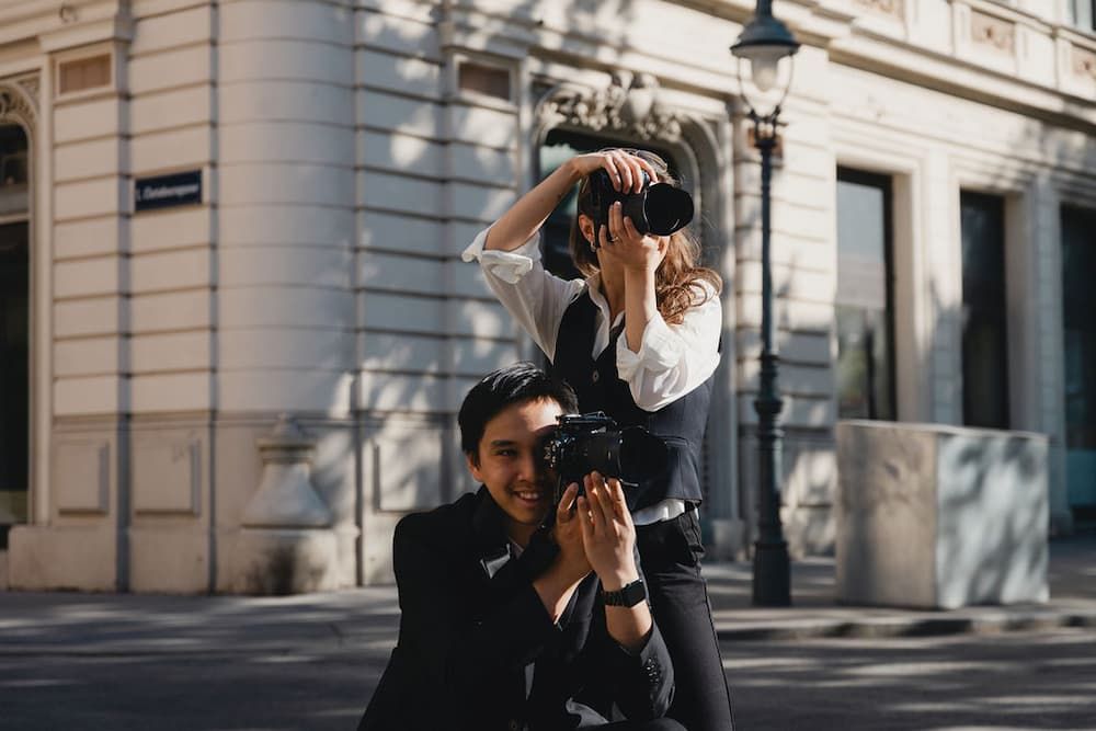 Two photographers capturing urban architecture on a sunny street, one crouching with a camera while the other shoots from above.