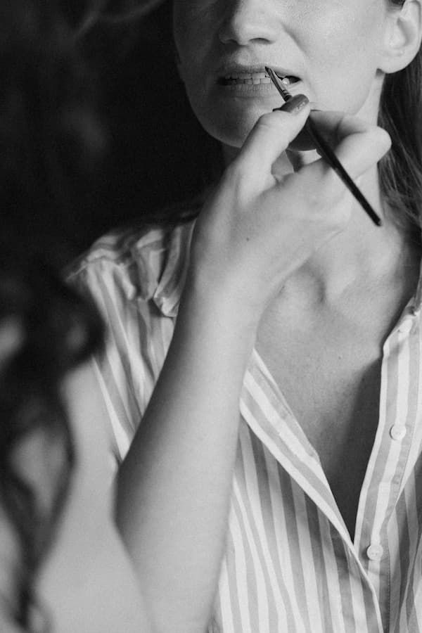 Black and white photo of a woman applying lipstick with a makeup brush, wearing a striped button-up shirt.