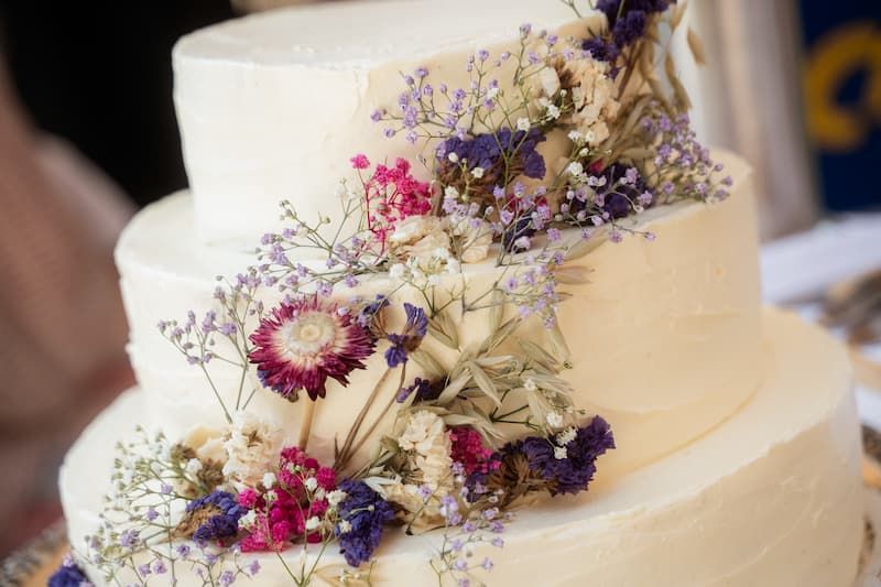 Elegant three-tier white wedding cake adorned with dried wildflowers in pink, purple, and cream cascading down the layers.