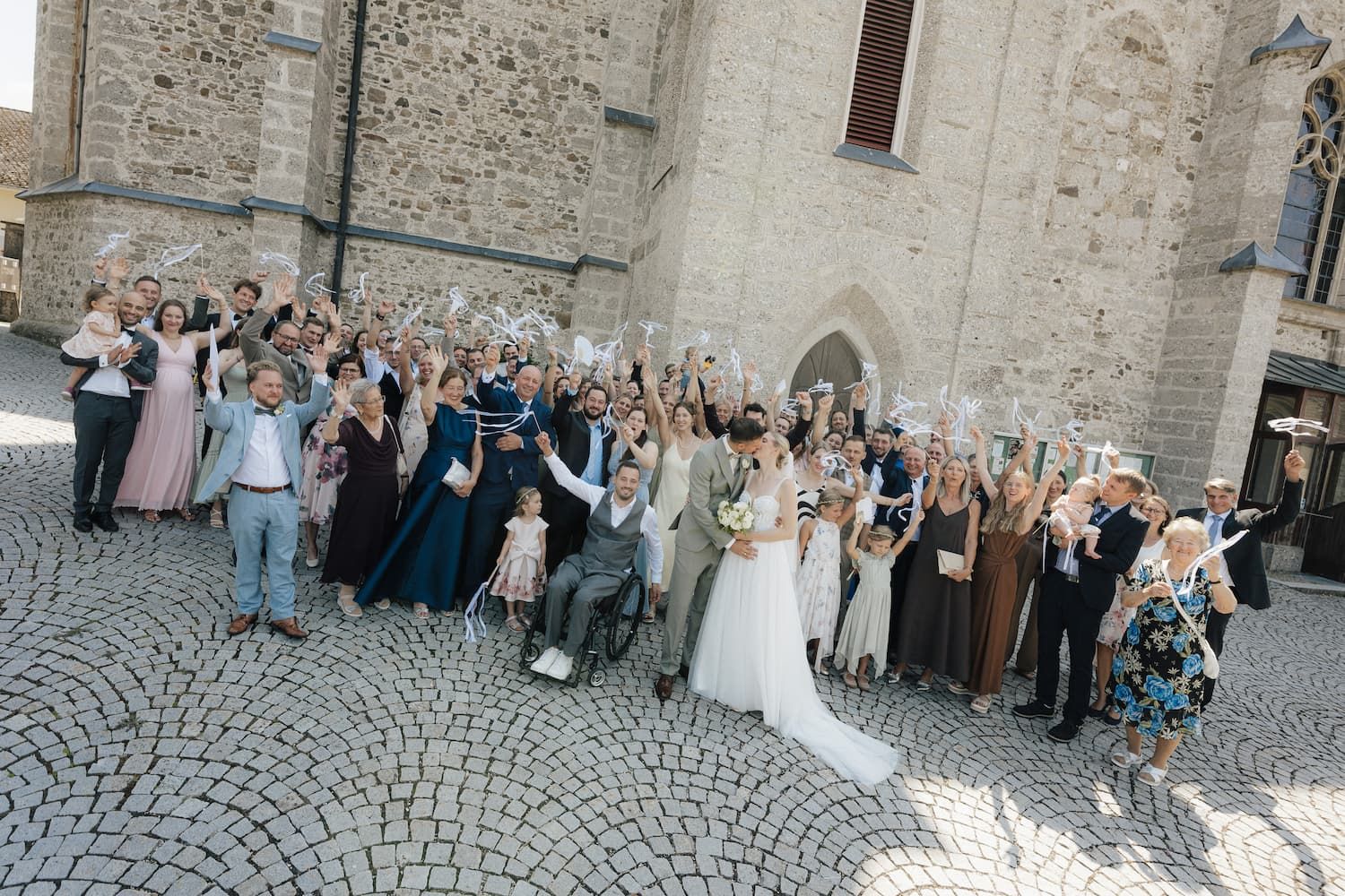 Large wedding group photo outside a stone church, bride and groom surrounded by guests waving white items in celebration.