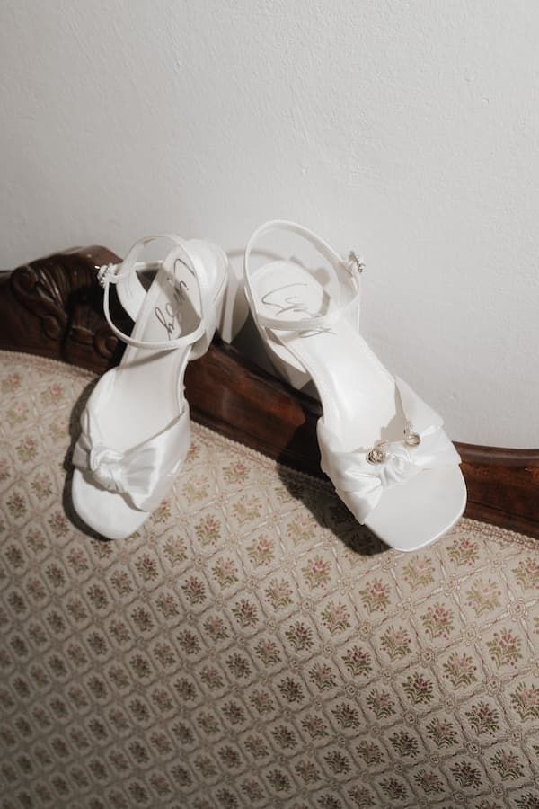 White bridal heeled sandals with ankle straps and bow detail resting on a vintage floral upholstered chair.