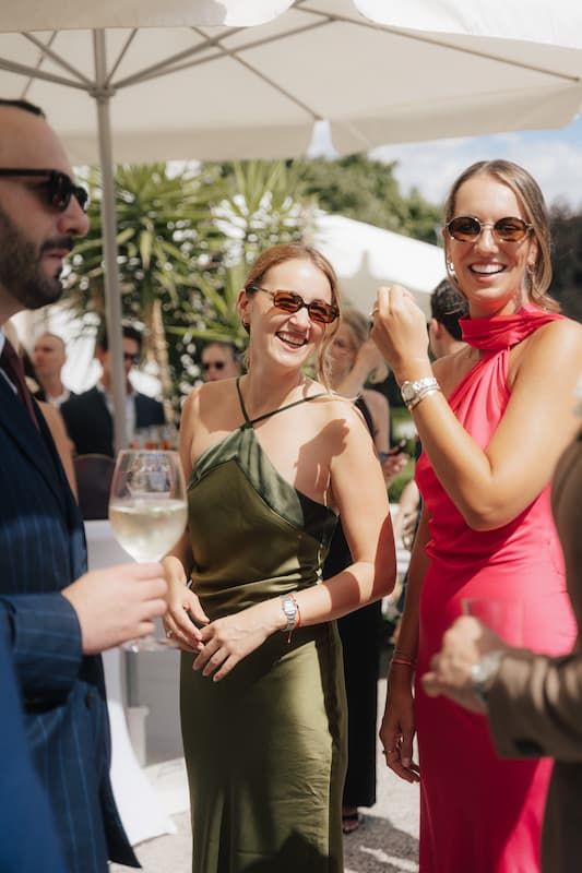 Two women in elegant green and red dresses laughing at an outdoor wedding reception with champagne glasses.
