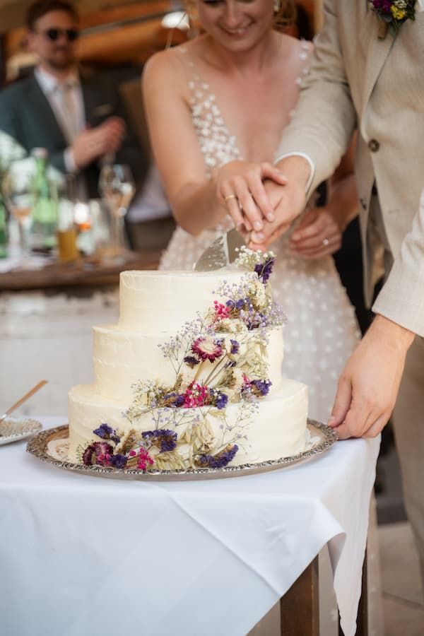 Bride and groom cutting a three-tier white wedding cake decorated with dried flowers on a silver tray.