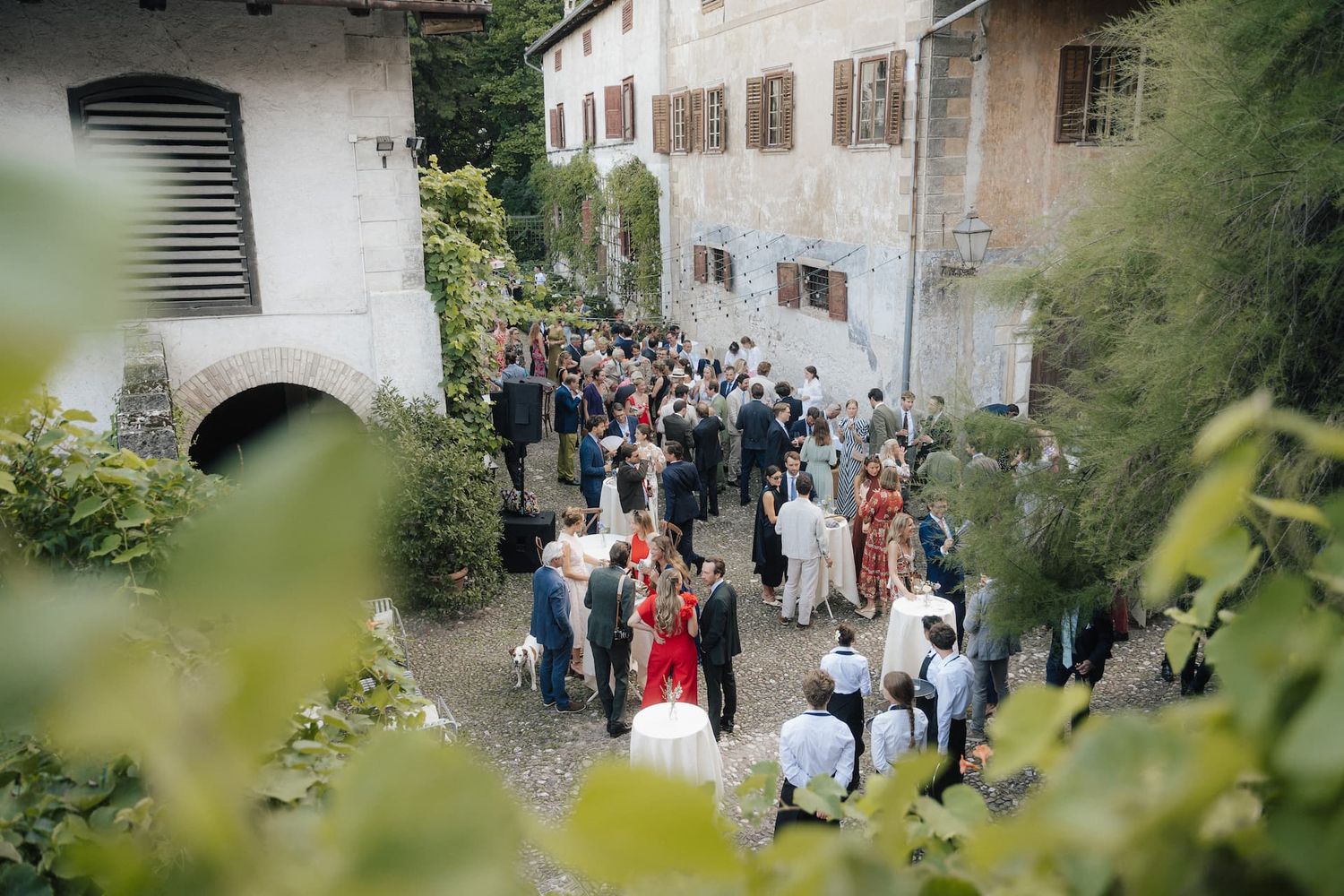 Elegant outdoor wedding cocktail hour in a rustic Italian courtyard with guests mingling among lush greenery.