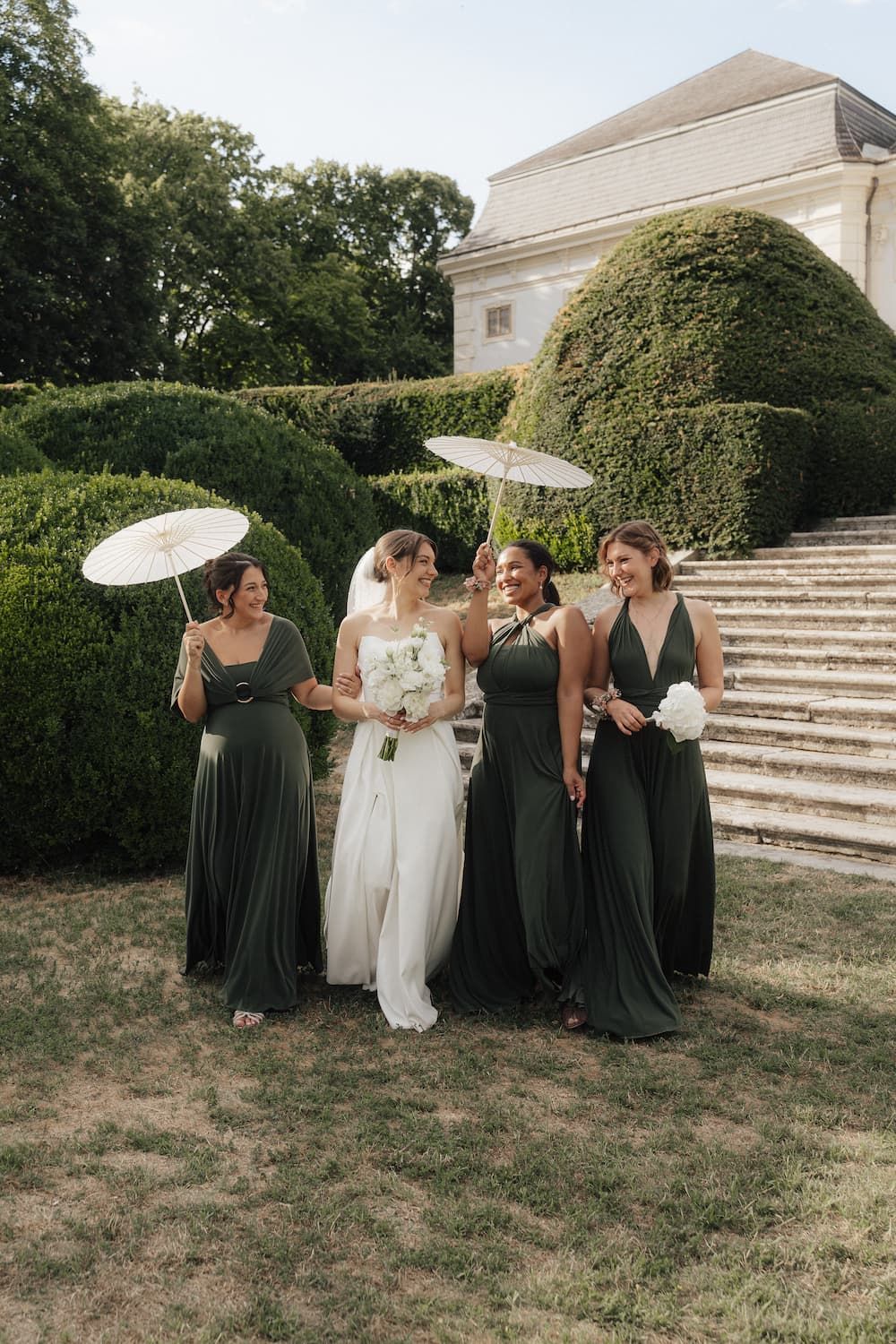 Bride in white gown with three bridesmaids in sage green dresses holding parasols in a garden.