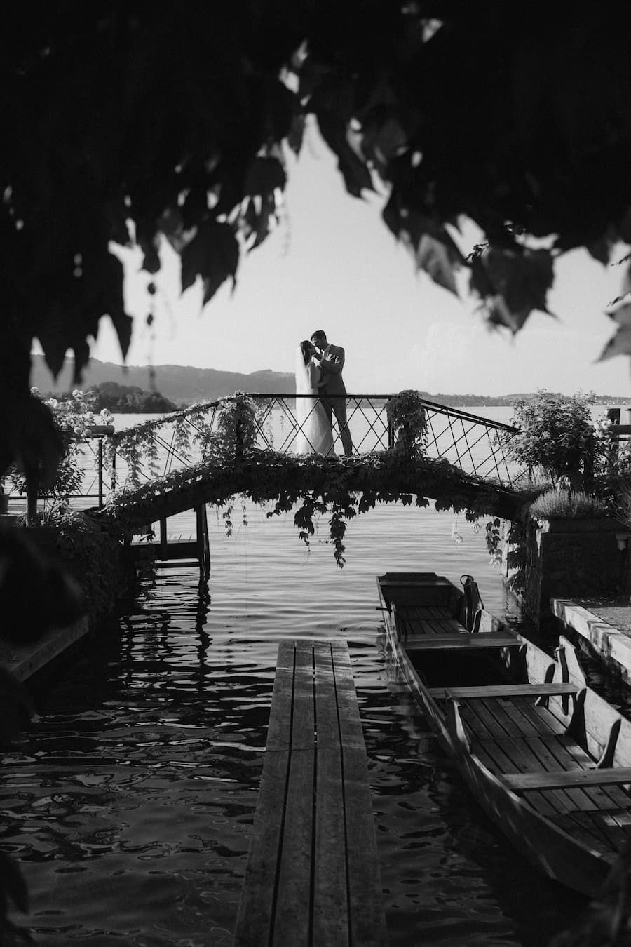 Black and white photo of a couple kissing on a decorated bridge over water, framed by lush leaves and boats.