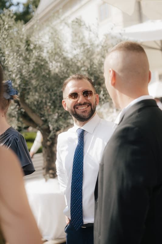 Smiling bearded man in white shirt and blue polka dot tie wearing sunglasses at an outdoor wedding reception.