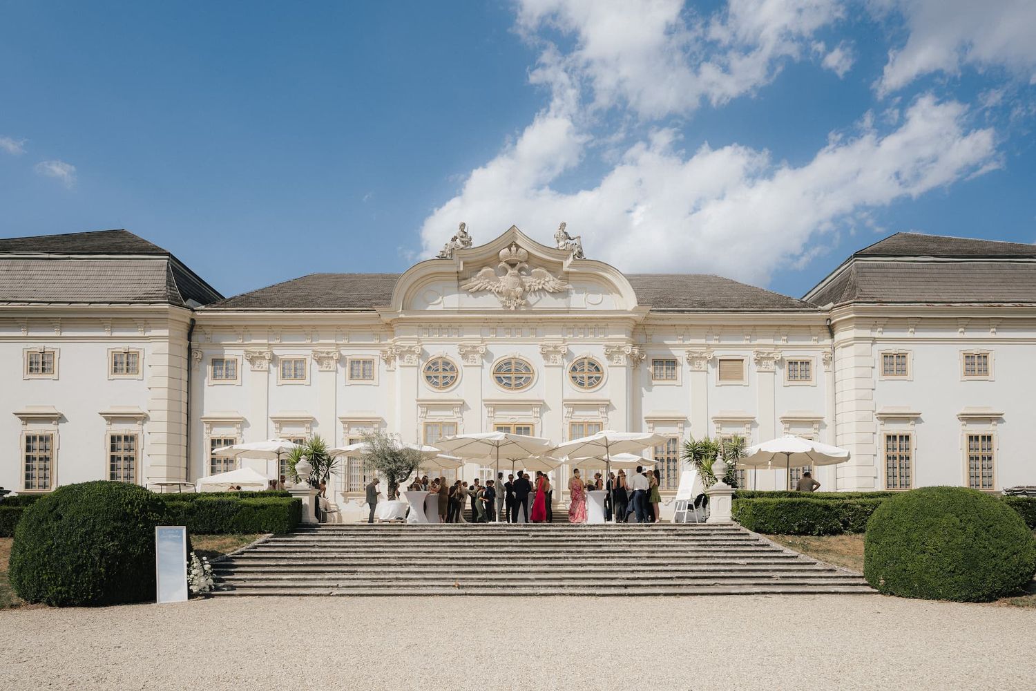 Elegant white baroque palace wedding venue with guests gathered on grand stone steps under a blue sky.