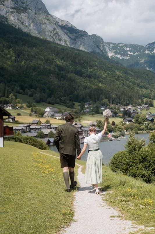 Couple in traditional Austrian attire walking hand-in-hand on a path with alpine village and mountain lake backdrop.