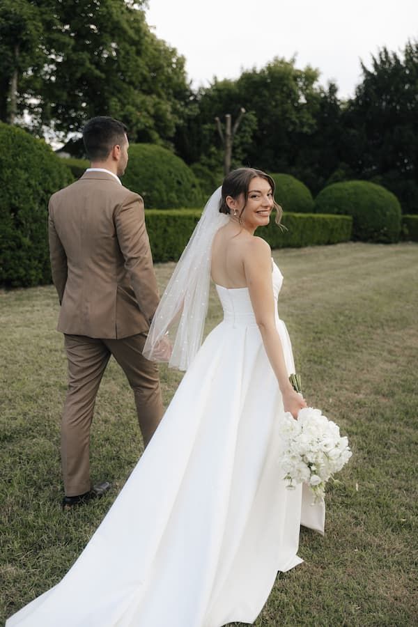 Bride in white gown with veil holds bouquet, smiling back at groom in tan suit walking through lush garden.