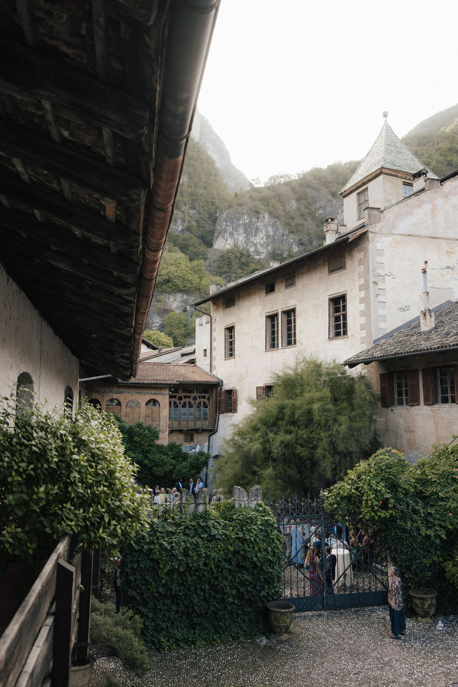 Historic alpine monastery courtyard with stone buildings, lush greenery, and mountain backdrop on a misty day.