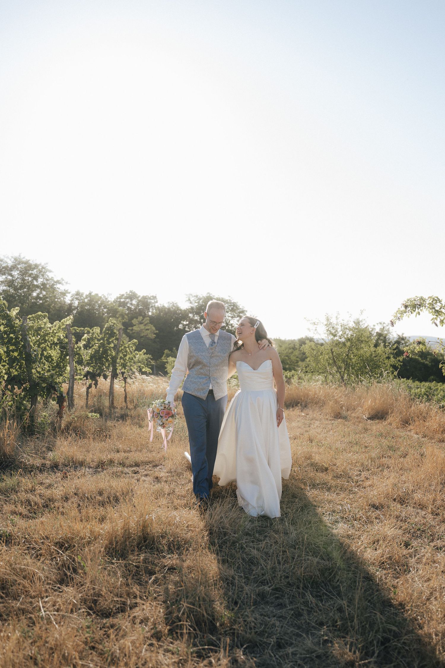 Bride and groom walking hand in hand through a sunlit field at golden hour on their wedding day.