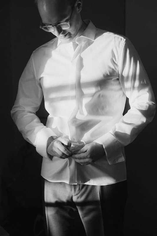 Black and white photo of a man in a white dress shirt buttoning up, illuminated by dramatic window light.