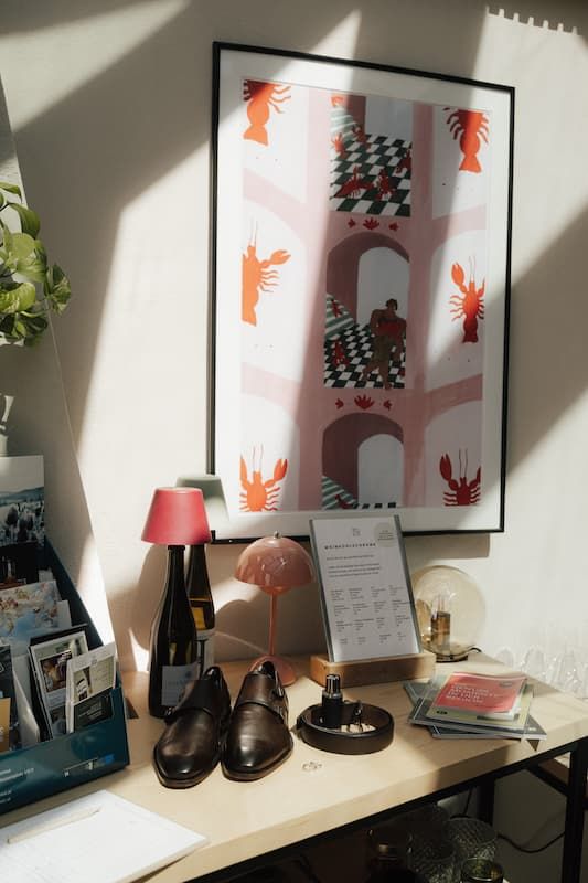 Desk display with brown leather shoes, pink lamps, and a framed lobster art print on a cream wall.