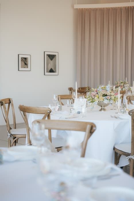Elegant wedding reception room with white tablecloths, wooden cross-back chairs, and pastel floral centerpieces in soft neutral tones.