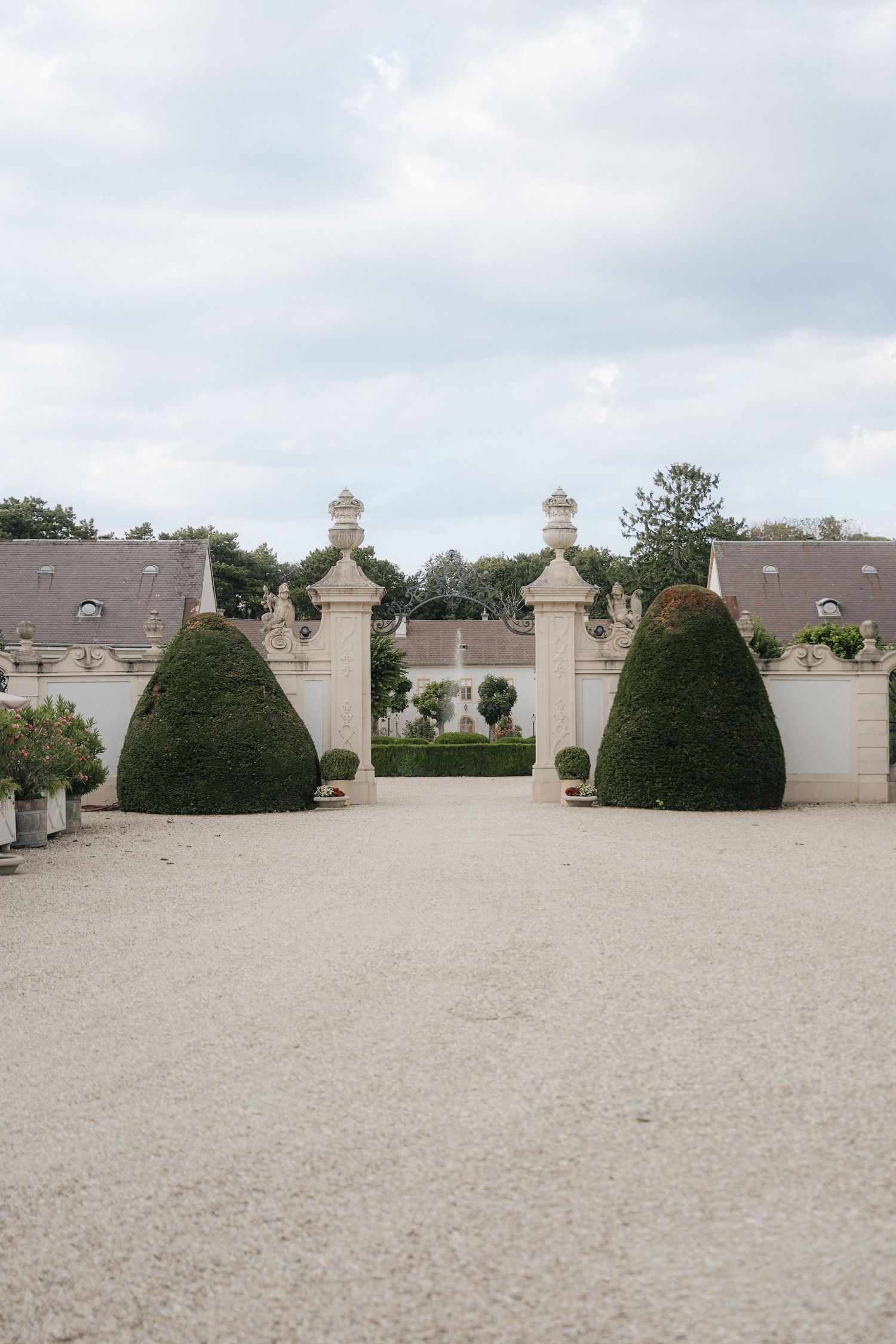 Elegant French château entrance with cone-shaped topiary bushes, stone pillars, and gravel courtyard under cloudy sky.