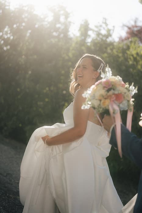 Radiant bride in white strapless gown holding colorful bouquet with pink ribbons, smiling joyfully in sunlit outdoor setting.