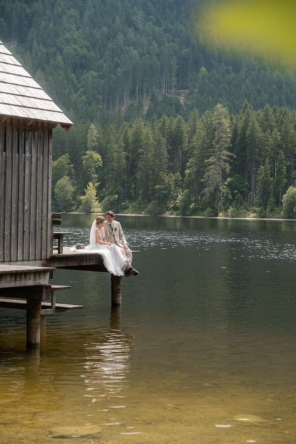 Bride and groom sitting on a wooden dock over a serene lake surrounded by lush green forest mountains.