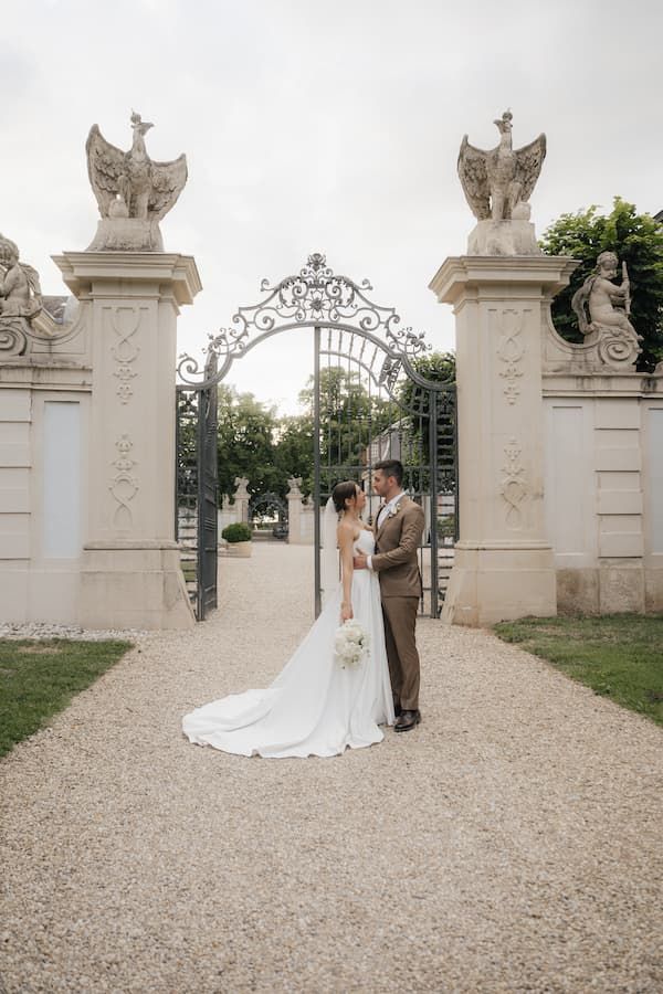 Bride and groom stand before ornate iron gates with stone eagle sculptures at an elegant European estate wedding venue.