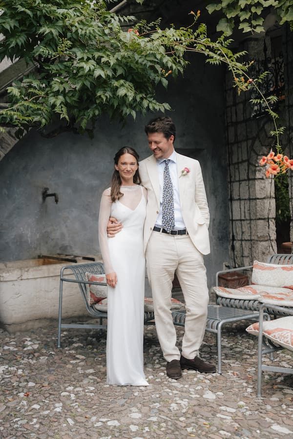 Bride in white gown and groom in cream suit pose together in a rustic outdoor courtyard setting.