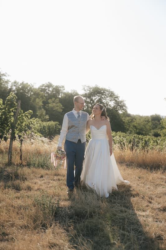 Bride and groom walking hand in hand through a sunlit vineyard field, smiling at each other on their wedding day.