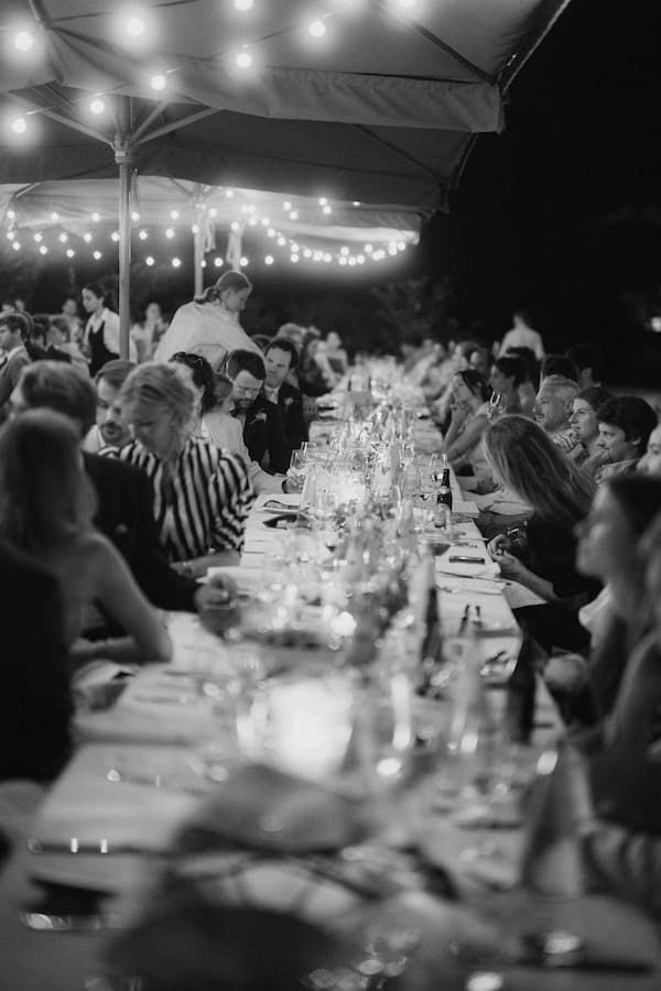 Black and white photo of elegant outdoor wedding reception dinner with string lights and long candlelit tables full of guests.