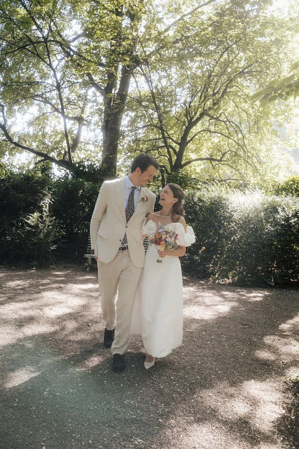A bride and groom walk together in a sunlit garden, she in a white dress holding a bouquet, he in a beige suit.