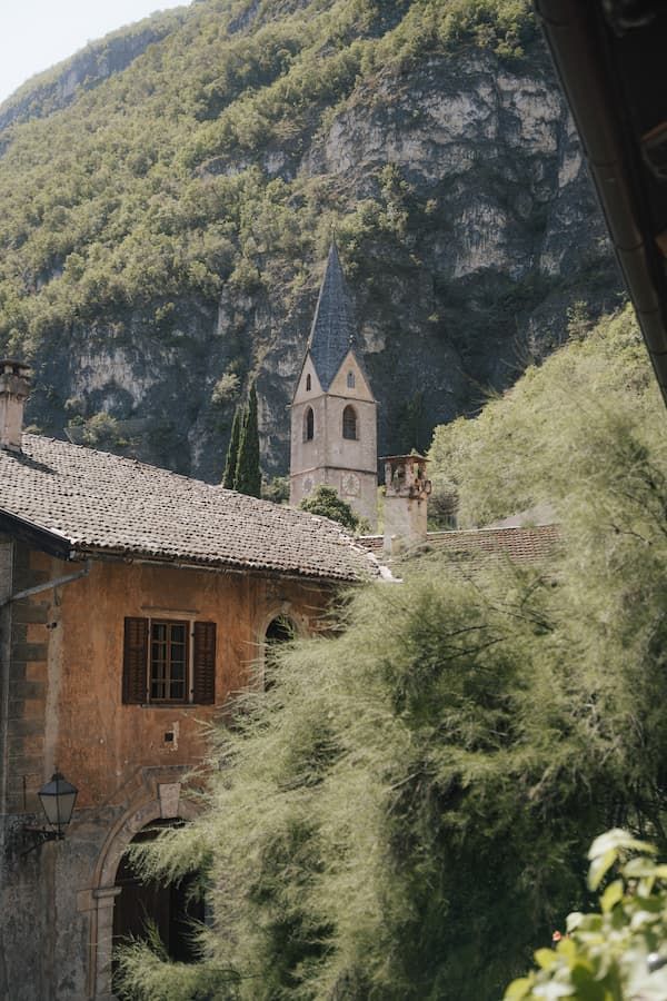Historic church tower rises above rustic stone buildings nestled among lush green trees with dramatic mountain cliffs in background.