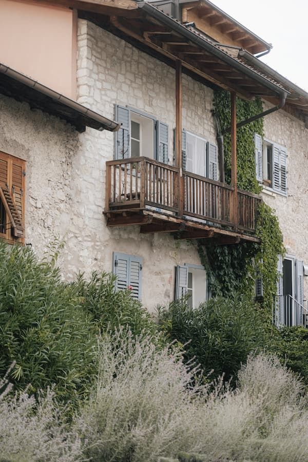 Stone building with wooden balcony, ivy, shuttered windows, and lush greenery in a charming European village setting.