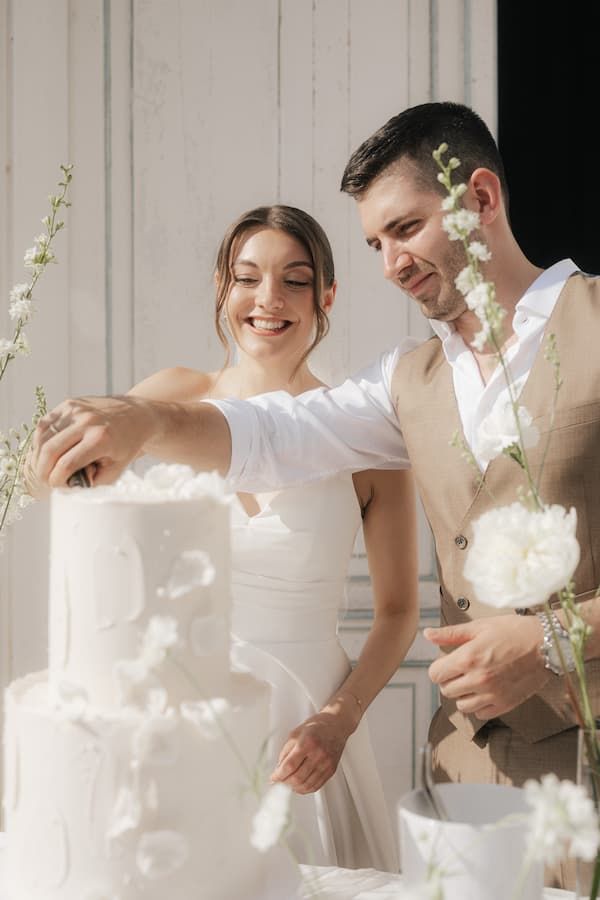 Smiling bride and groom cutting a elegant white tiered wedding cake decorated with flowers.