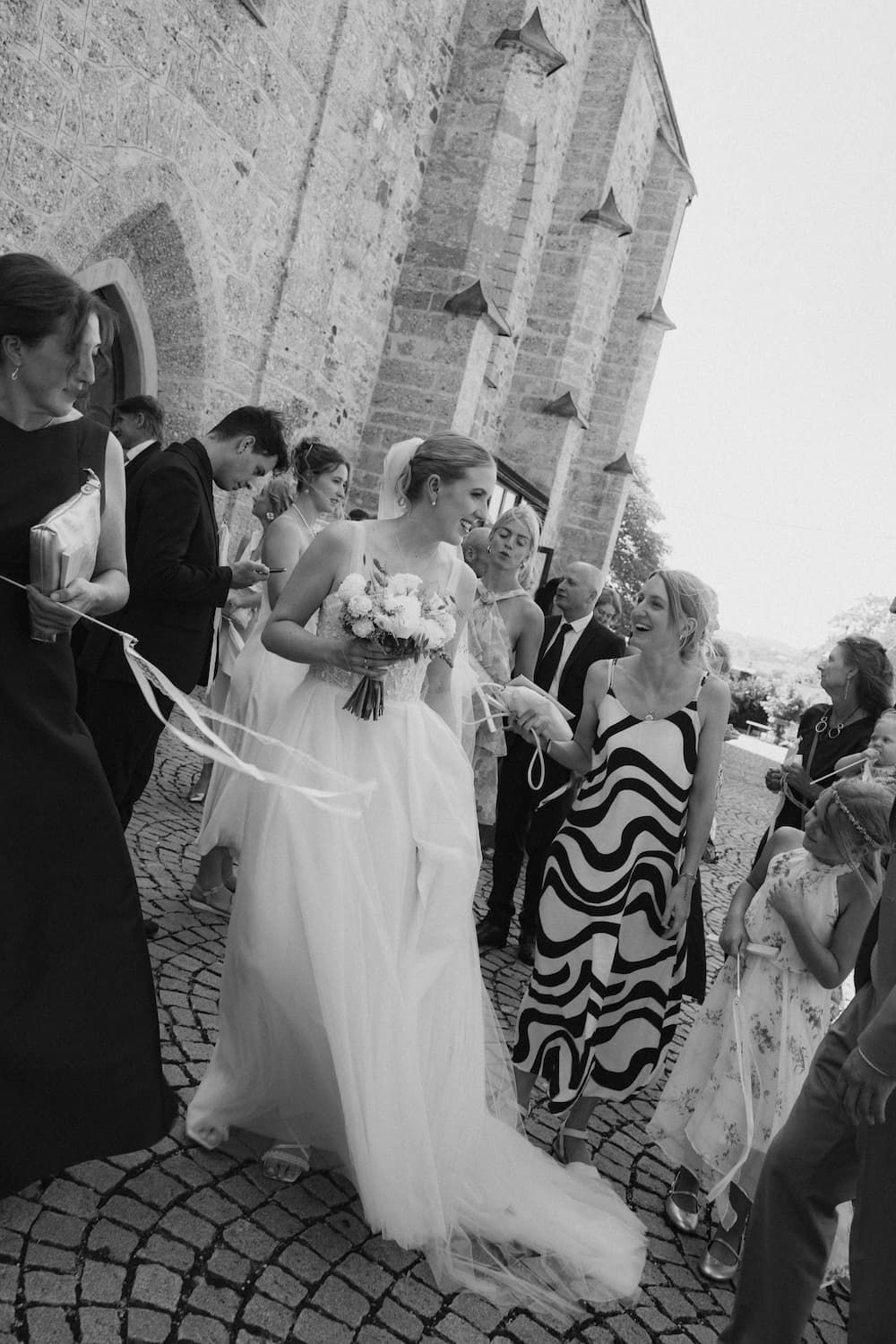 Black and white photo of a bride in a white gown holding a bouquet, surrounded by guests outside a stone church.