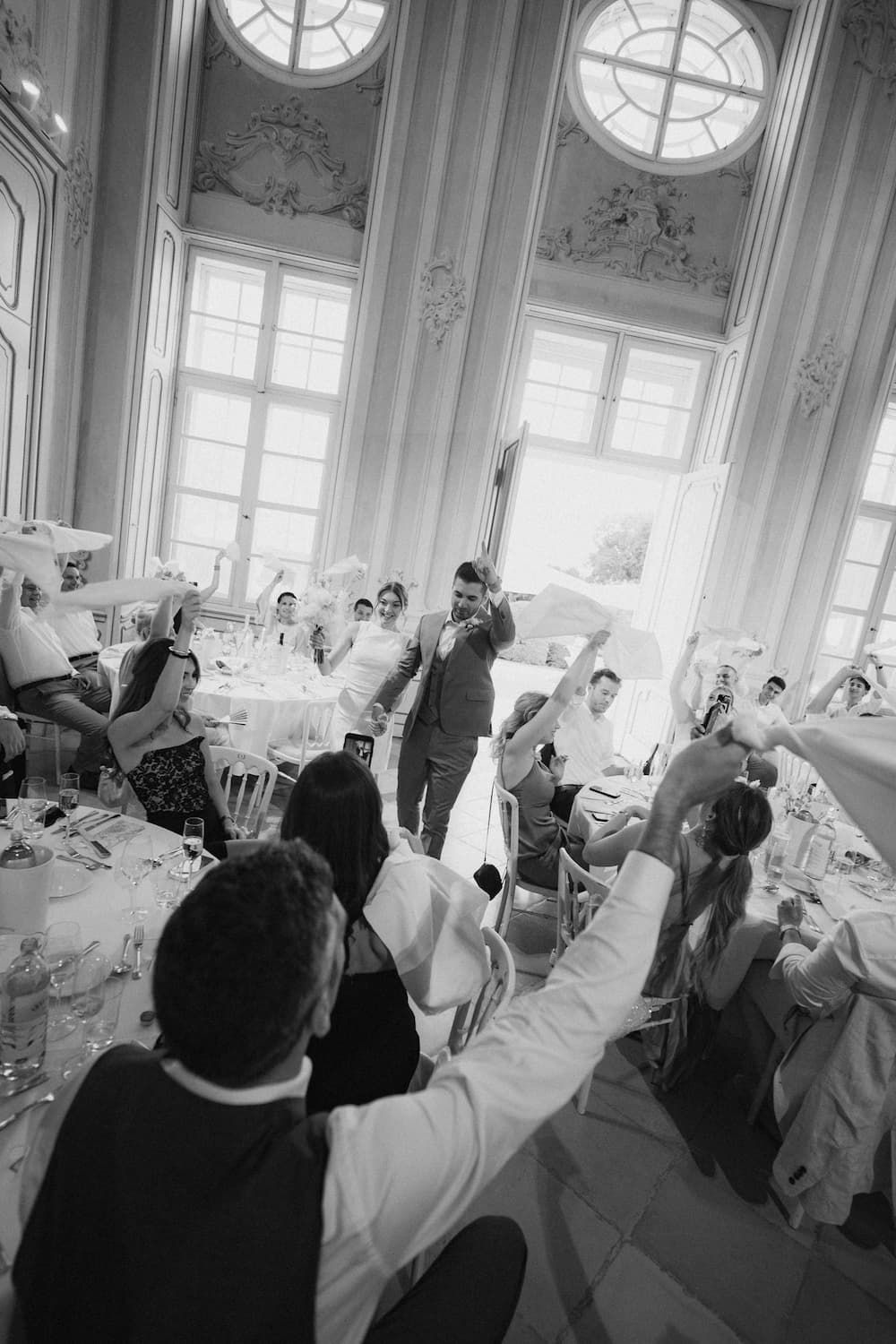 Black and white photo of wedding guests raising hands in elegant ballroom with tall columns and ornate decor.