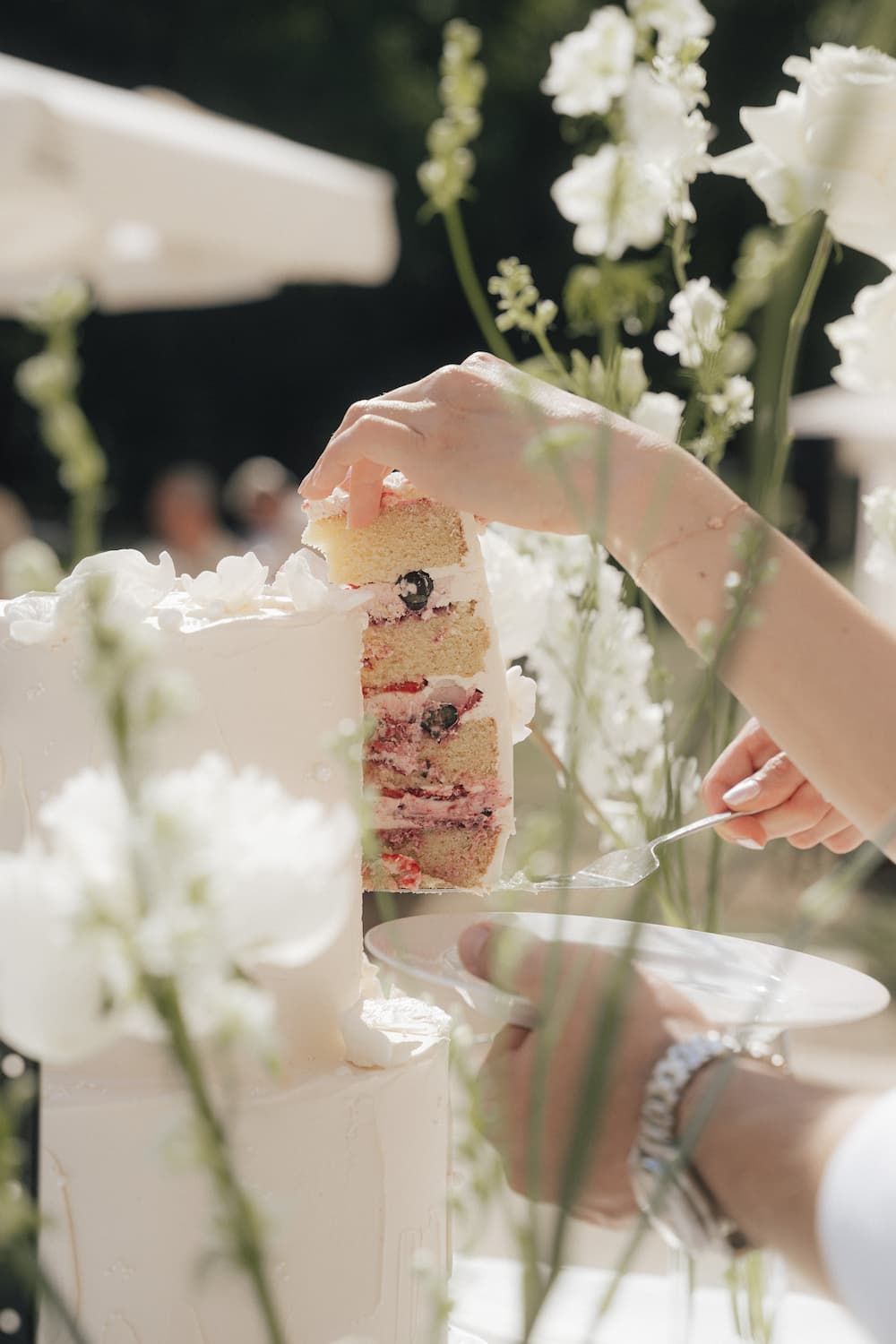 Hands cutting a white wedding cake revealing colorful berry layers, surrounded by white flowers outdoors.