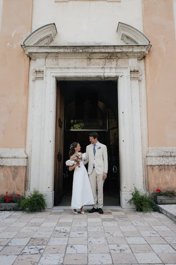 Bride and groom stand together in doorway of elegant European church, bride holding bouquet in white dress, groom in beige suit.