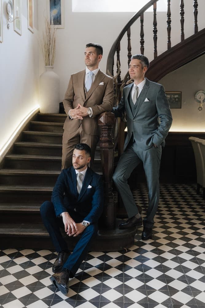 Three men in elegant tailored suits pose on a classic checkered floor staircase, wearing brown, grey, and navy ensembles.