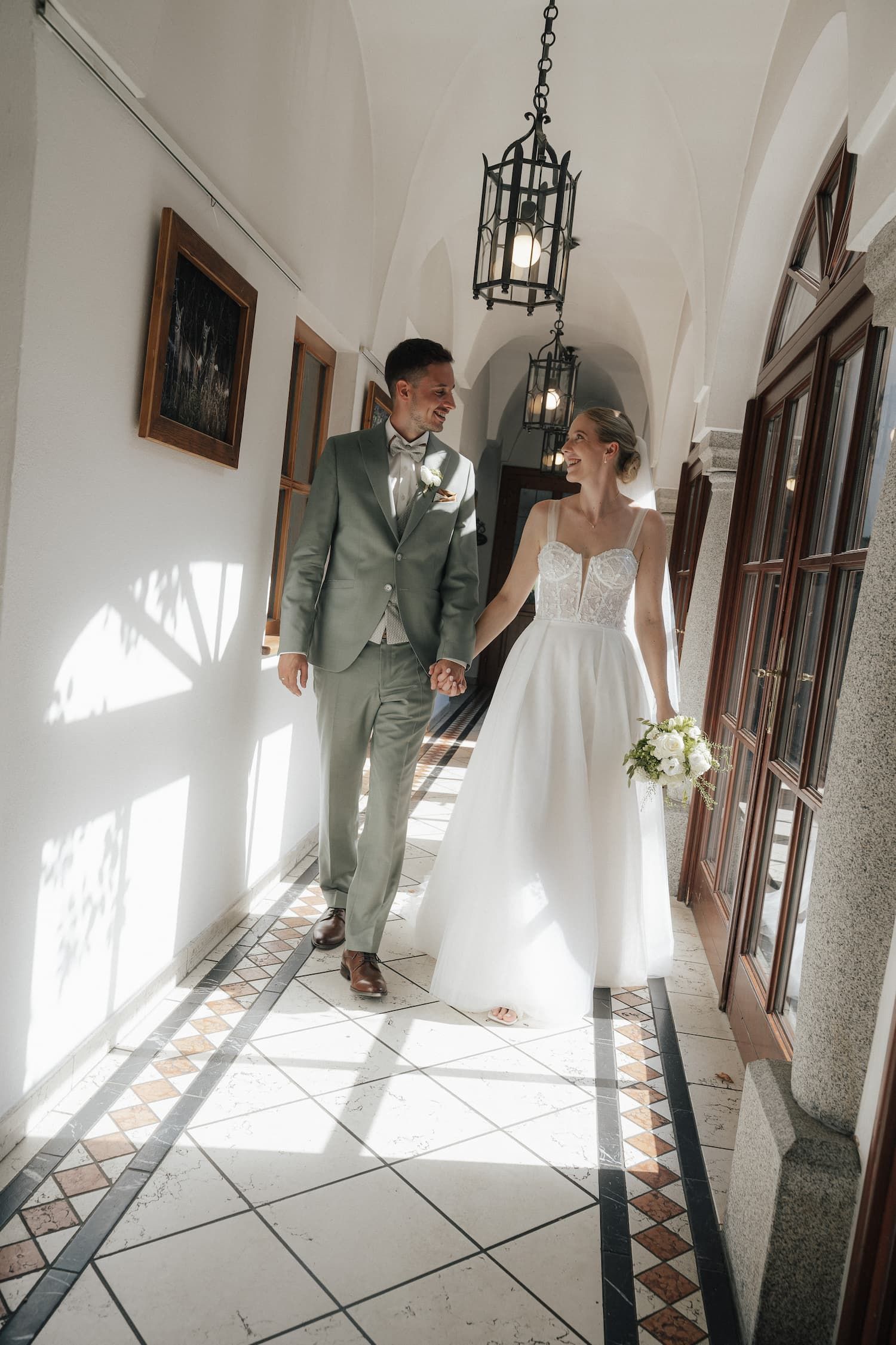 Bride and groom walk hand in hand through sunlit corridor with arched ceiling, tile floors, and hanging lanterns.